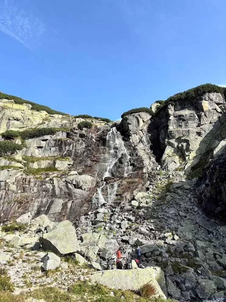 A tall waterfall falling down granite slabs in the High Tatras, two walkers resting on a boulder below it.