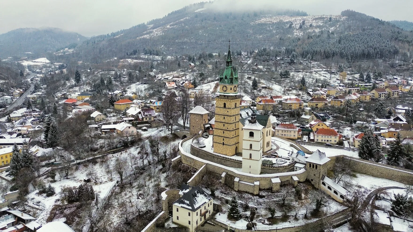 Kremnica Castle from the air in winter: the fortified precinct inside its curved stone wall, the tall green-spired tower of the Church of St Catherine rising from the middle, with the snow-covered roofs of the town and the wooded hills around it.