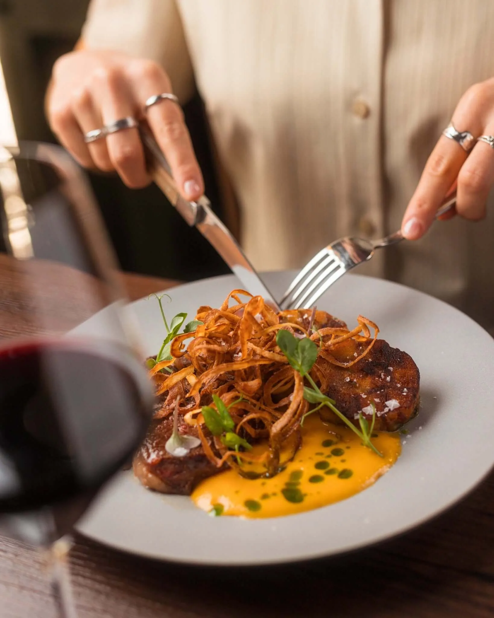 A main course being cut at Urban Betyár in Budapest: grilled meat under a nest of crisp fried onions, on a bright orange purée dotted with herb oil and micro-herbs, with a glass of red wine on the dark wooden table.