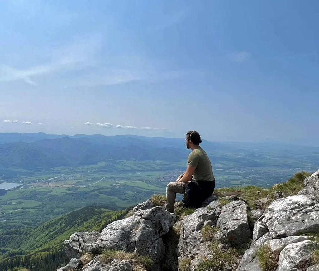 A walker sitting on the limestone summit rocks in the Malá Fatra, looking out over the valley and the hills beyond.