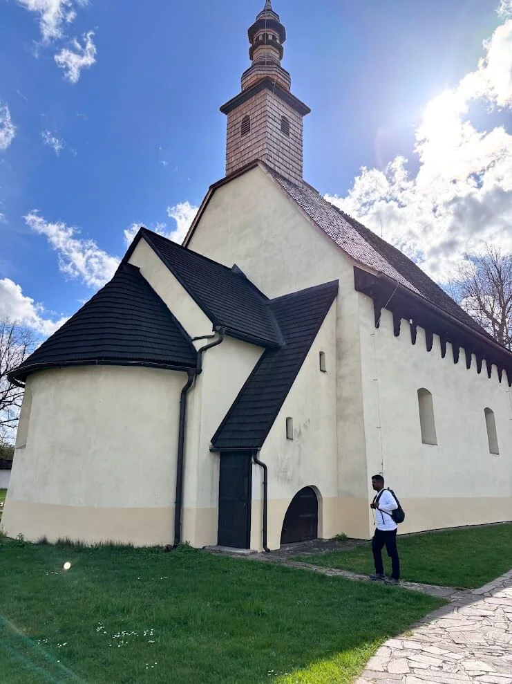A white medieval church with a tall shingled tower and steep dark roof standing on the grass, a visitor looking up at it.