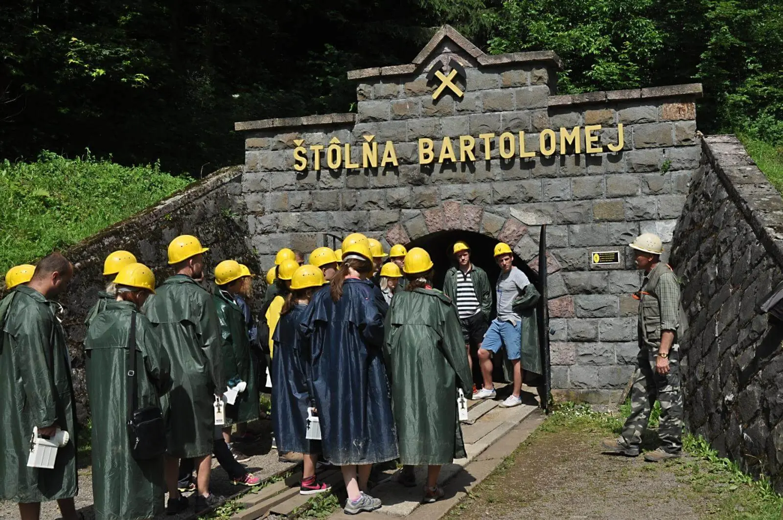 A group in yellow miners’ helmets and waterproof capes, carrying lamps, waiting with their guide at the stone portal of the Štôlňa Bartolomej adit at the open-air mining museum in Banská Štiavnica, the crossed hammers of the miners’ emblem above the entrance.