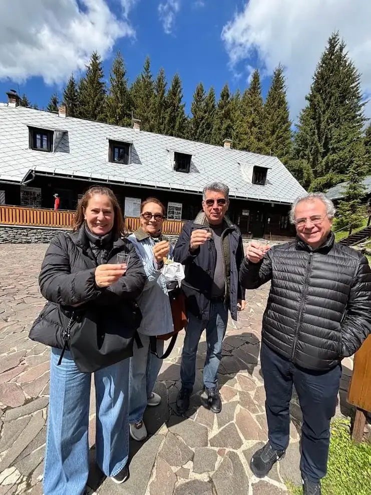 Four guests raising small glasses in a toast outside a wooden mountain chalet, spruce forest behind.