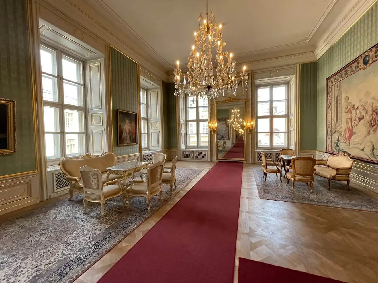 A salon inside the Primate’s Palace in Bratislava: green silk walls hung with an English tapestry, gilded furniture on patterned carpets, a crystal chandelier overhead and the enfilade of rooms running away through a mirrored door.