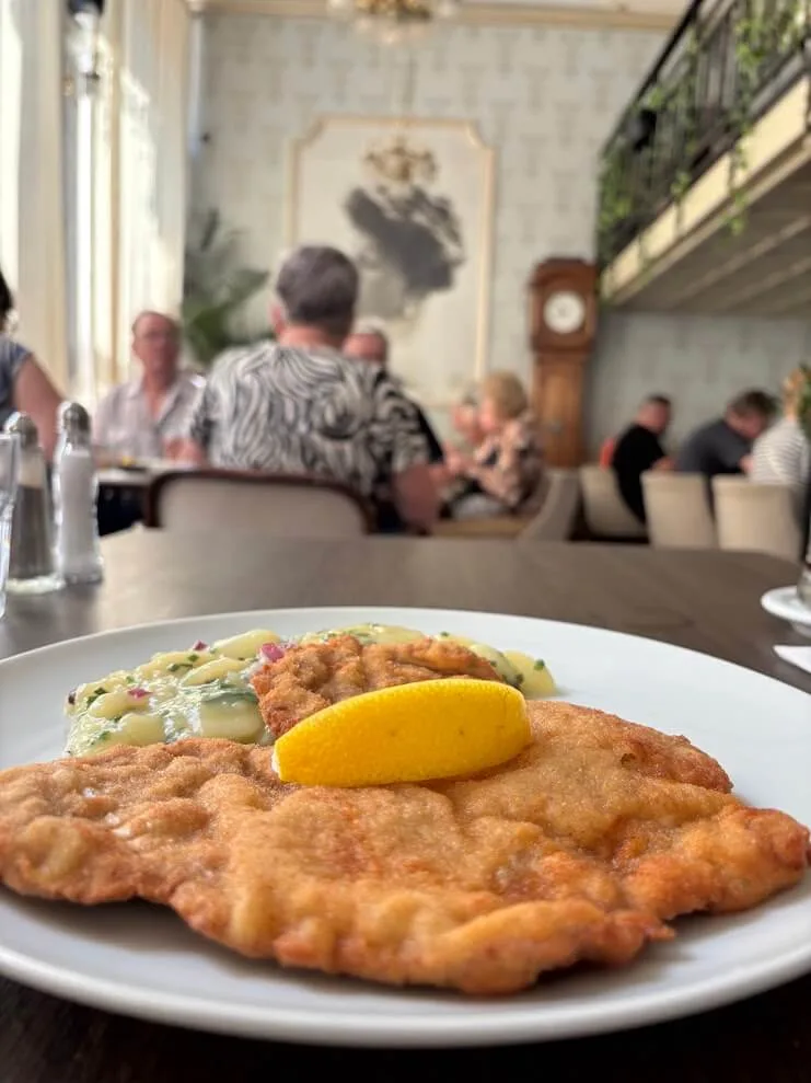 A breaded schnitzel with potato salad and a wedge of lemon on a white plate at Café Štefánka in Bratislava, with guests at the tables and the grandfather clock of the panelled dining room behind.