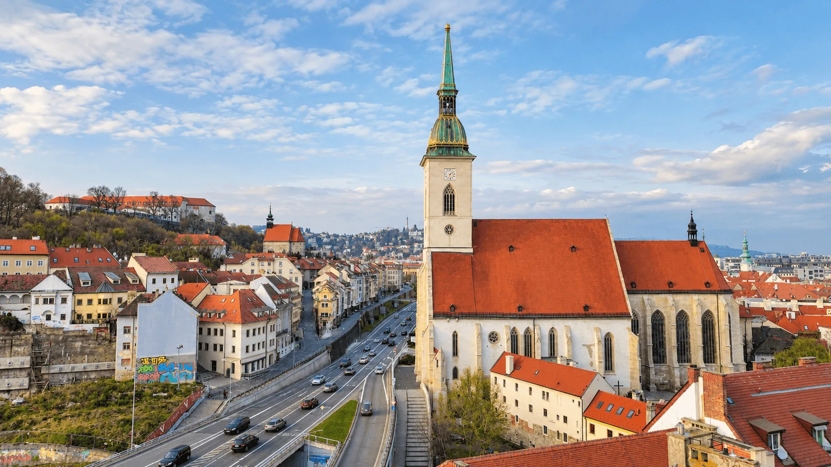 St Martin’s Cathedral in Bratislava seen from above: the Gothic nave under its red roof and the tower with its green copper spire, the old town rooftops on one side and Bratislava Castle on its hill behind.
