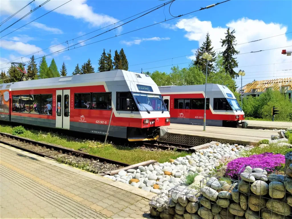 Two red and white Tatra Electric Railway trains waiting at a station platform in the High Tatras, spruce forest behind.