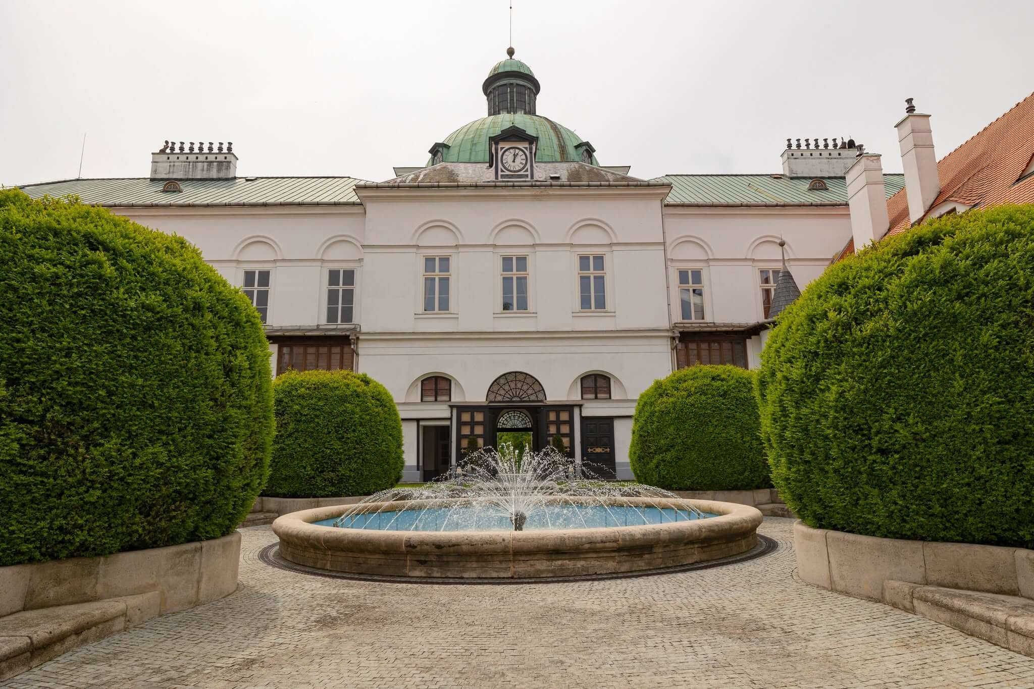 Topoľčianky Castle, the white manor that served as the summer residence of the first Czechoslovak president, its green copper clock dome rising beyond the clipped topiary and the fountain in the cobbled courtyard.