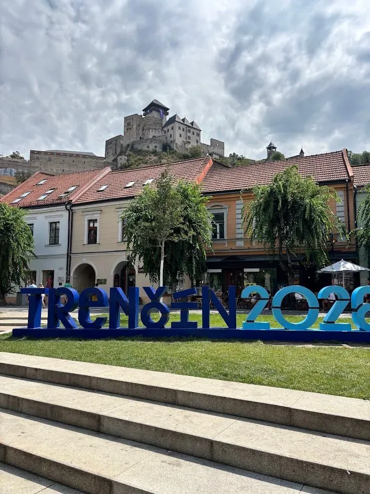 Trenčín’s main square with the plague column and the TRENČÍN 2026 sign, and Trenčín Castle on the rock above the rooftops.