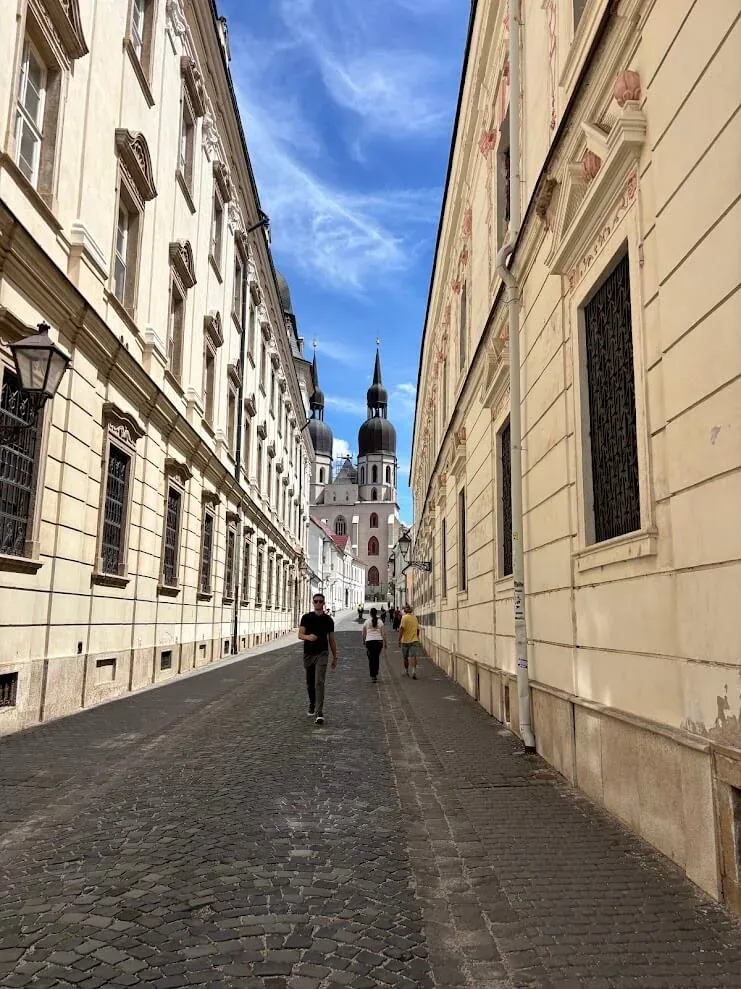 A narrow cobbled street between tall façades in Trnava’s old town, leading to the twin towers of St John the Baptist Cathedral at the far end.