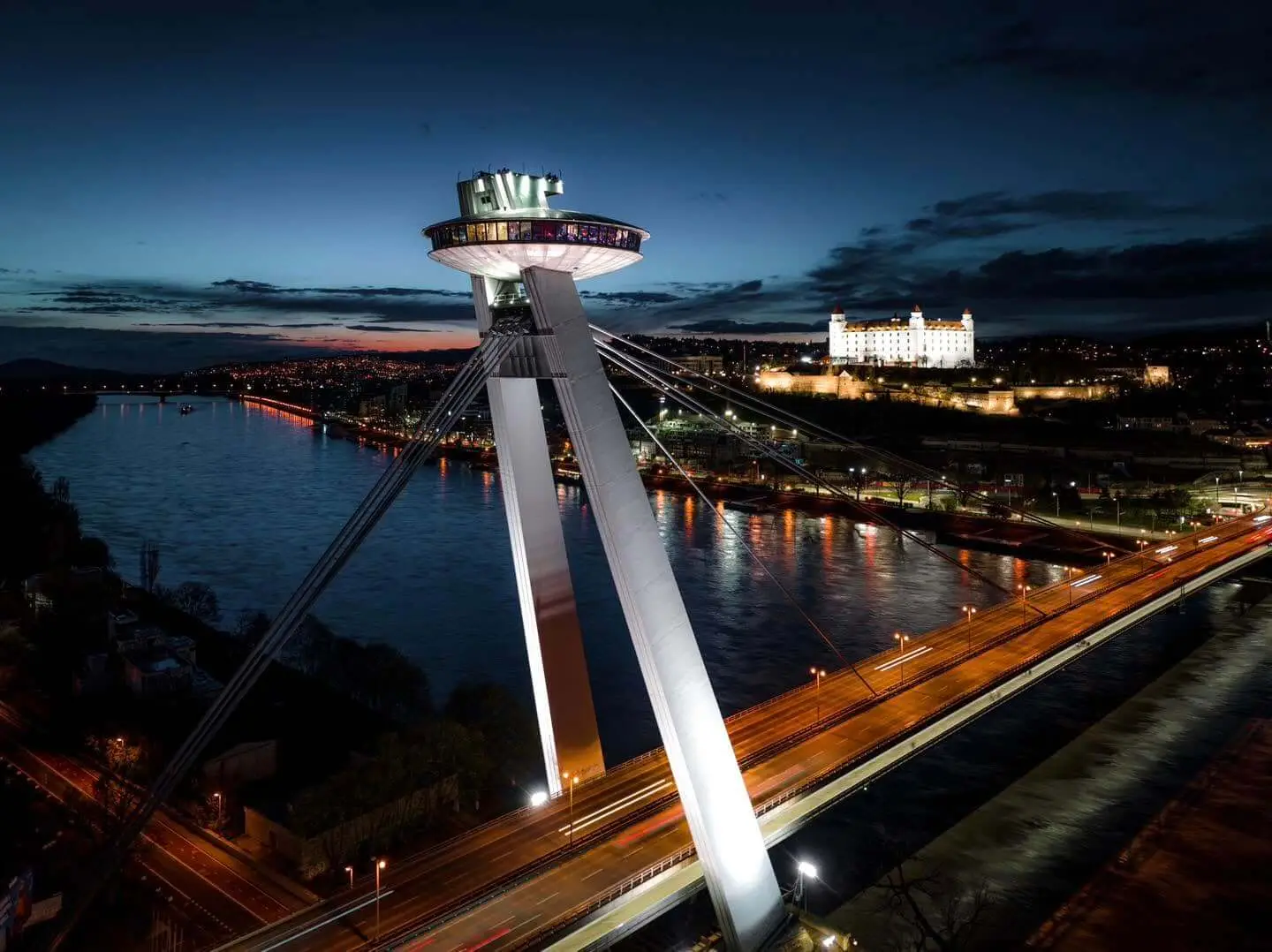 The UFO observation deck lit up on top of the pylon of Most SNP in Bratislava after dark, traffic streaming across the bridge beside the Danube, with Bratislava Castle floodlit on its hill across the river.