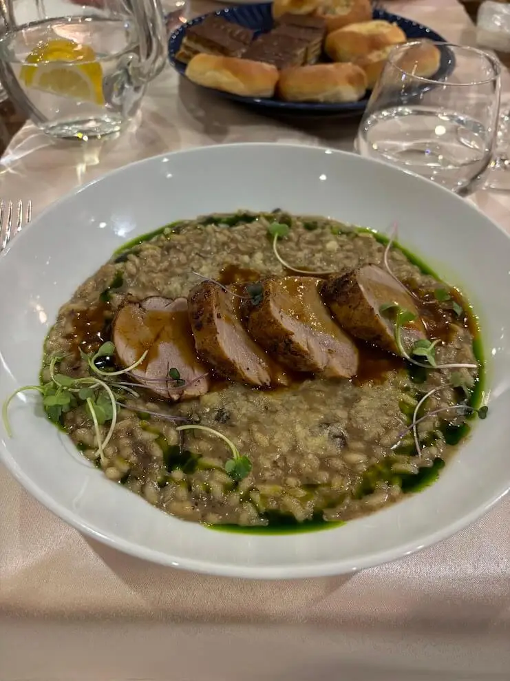 A main course served at a group wine dinner: slices of roast pork tenderloin in a dark jus on barley and mushroom risotto, dressed with micro-herbs and herb oil, with bread rolls and layered dessert slices on the table behind.