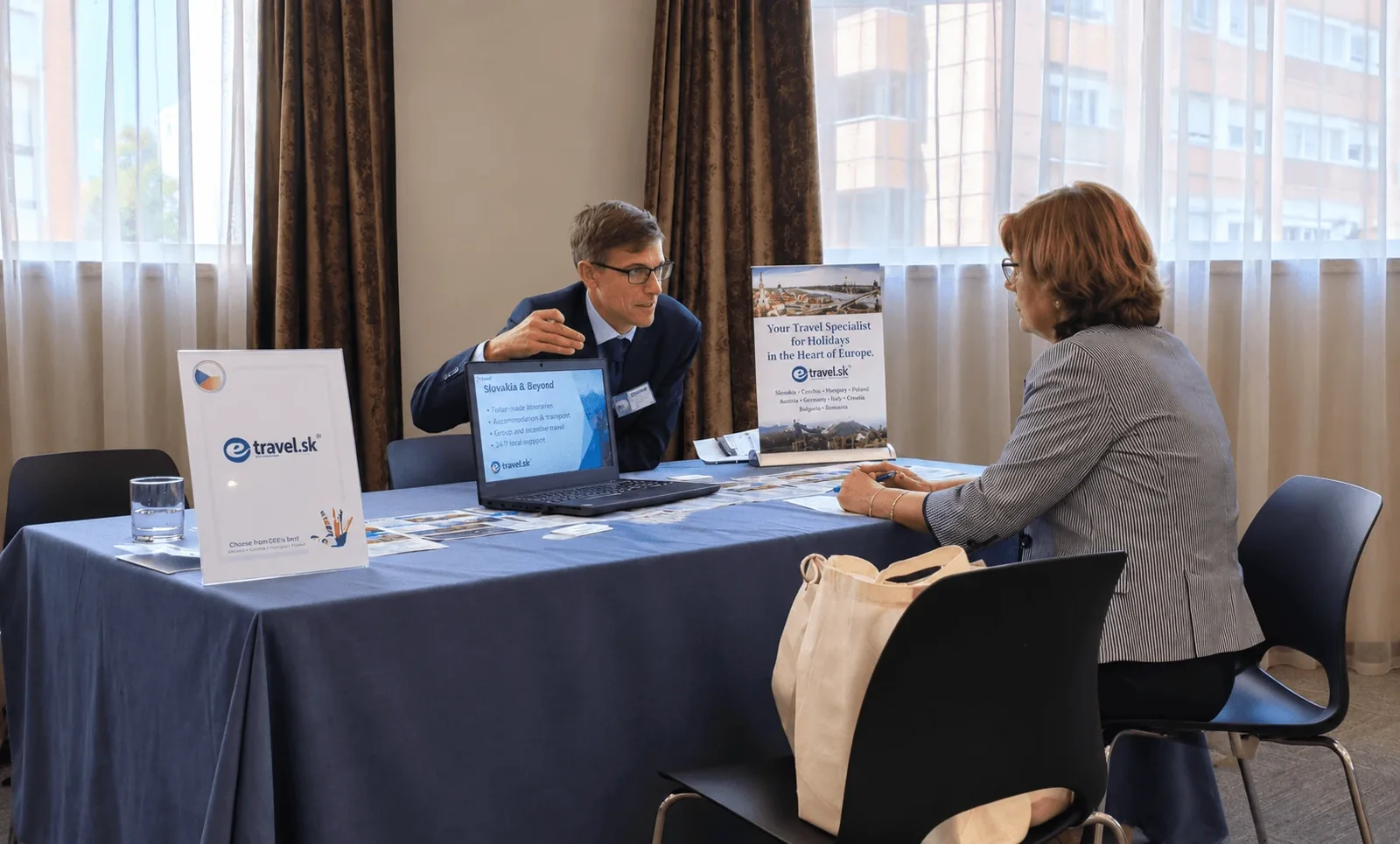 Un representante de e-travel.sk conversando con una compradora al otro lado de la mesa en un workshop profesional, con los expositores de Eslovaquia de la empresa y un portátil entre ambos.