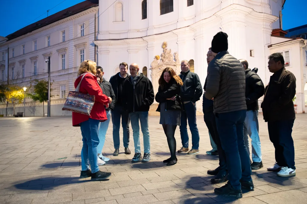 A guide with a corporate group outside the Trinitarian Church — the Church of St John of Matha — on Župné námestie in Bratislava, its curved white baroque façade and gilded portal sculpture lit by low sun.