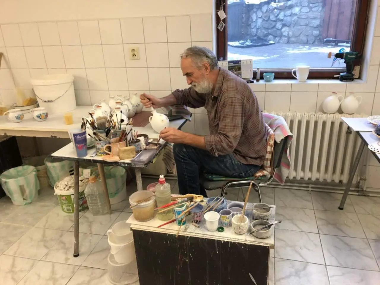 A craftsman hand-painting a majolica jug at his workbench in a pottery workshop in Pezinok, brushes and glaze pots spread around him.