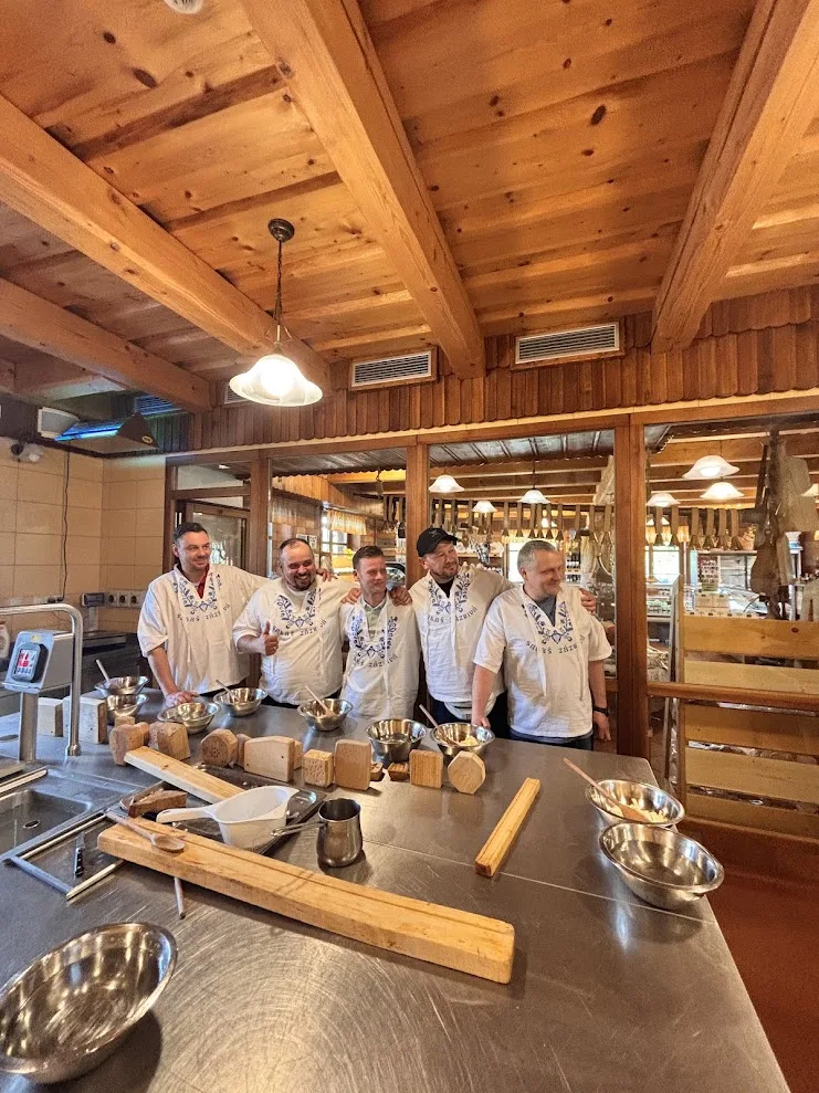 Five participants in embroidered Slovak aprons behind the steel counter at a sheep’s-cheese workshop in Zázrivá near Žilina, the wooden moulds and paddles laid out in front of them.