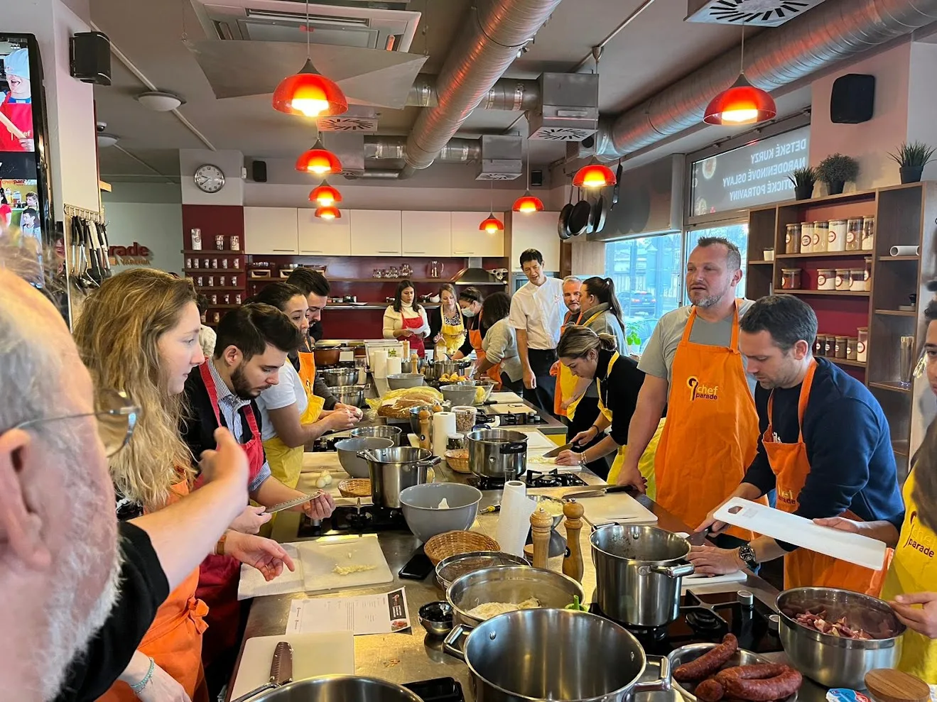 A team cooking class in progress at the Chefparade cooking school in Bratislava, delegates in orange and yellow aprons preparing food along a shared steel counter.