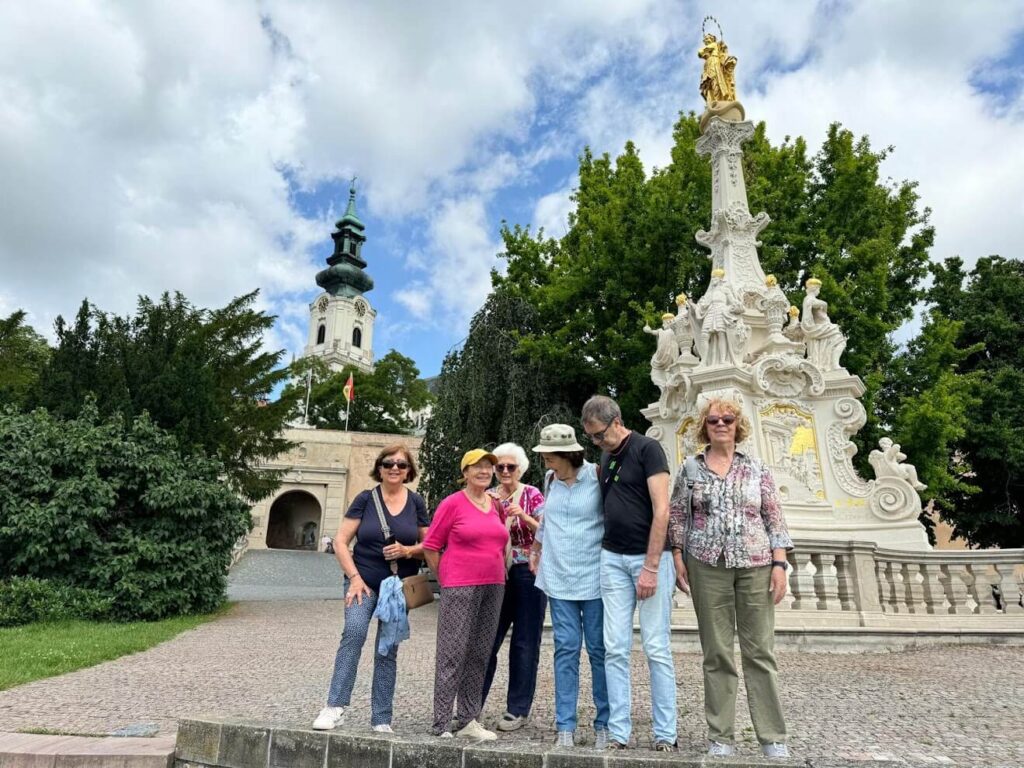 A group of tourists standing on a cobblestone plaza in Nitra during Central European Itinerary, Slovakia, next to an ornate white Baroque monument with gold accents topped with a religious statue. In the background is a church tower with a green spire and an archway entrance to what appears to be a castle or historic complex, all under a partly cloudy sky.