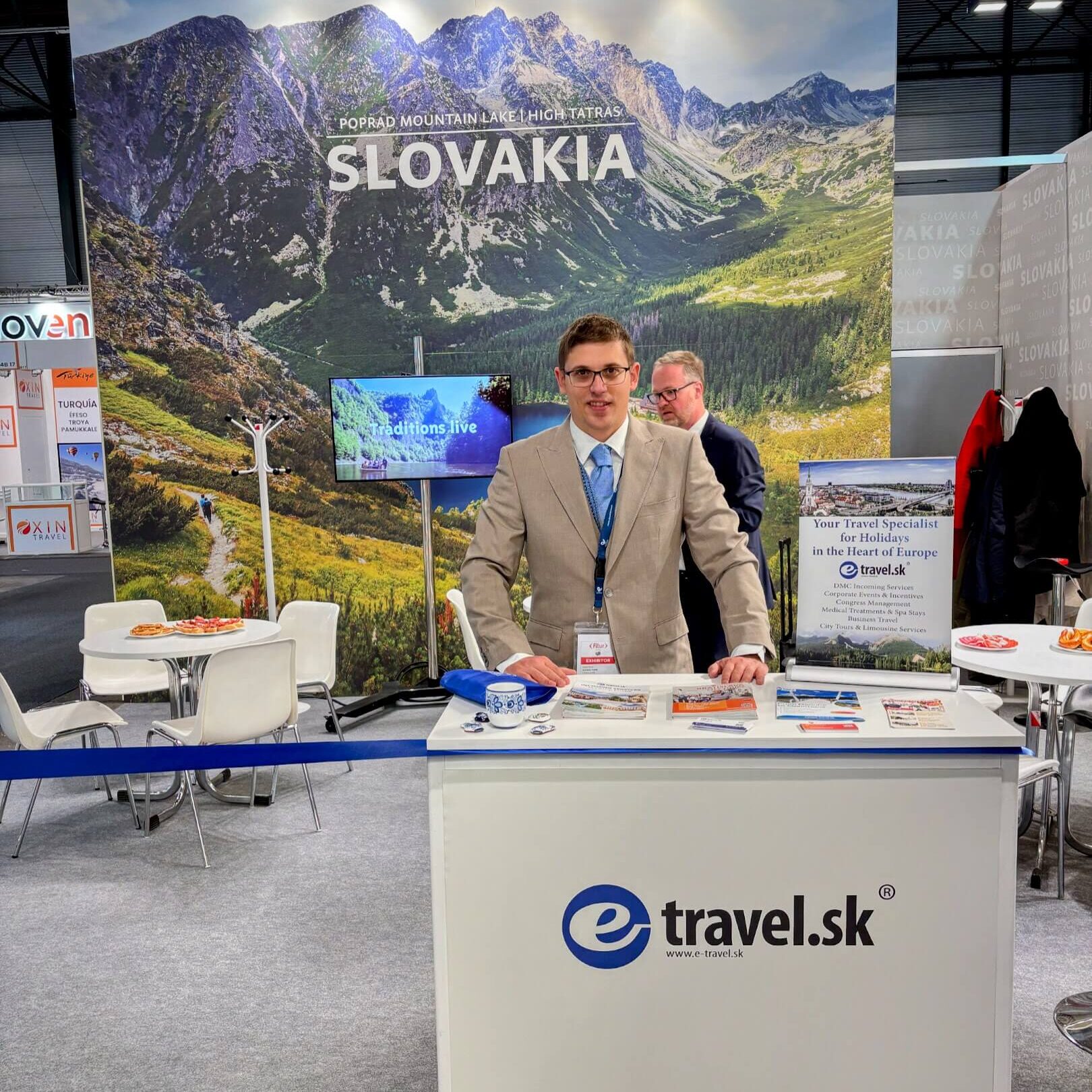 A man in a beige suit and blue tie stands behind a promotional booth for e-travel.sk at the FITUR travel trade fair. The booth features brochures, souvenirs, and a banner promoting Slovakia as a travel destination. Behind him, a large backdrop displays an image of the High Tatras mountains with the text "SLOVAKIA" and "Poprad Mountain Lake - High Tatras." Other exhibitors and visitors are visible in the background