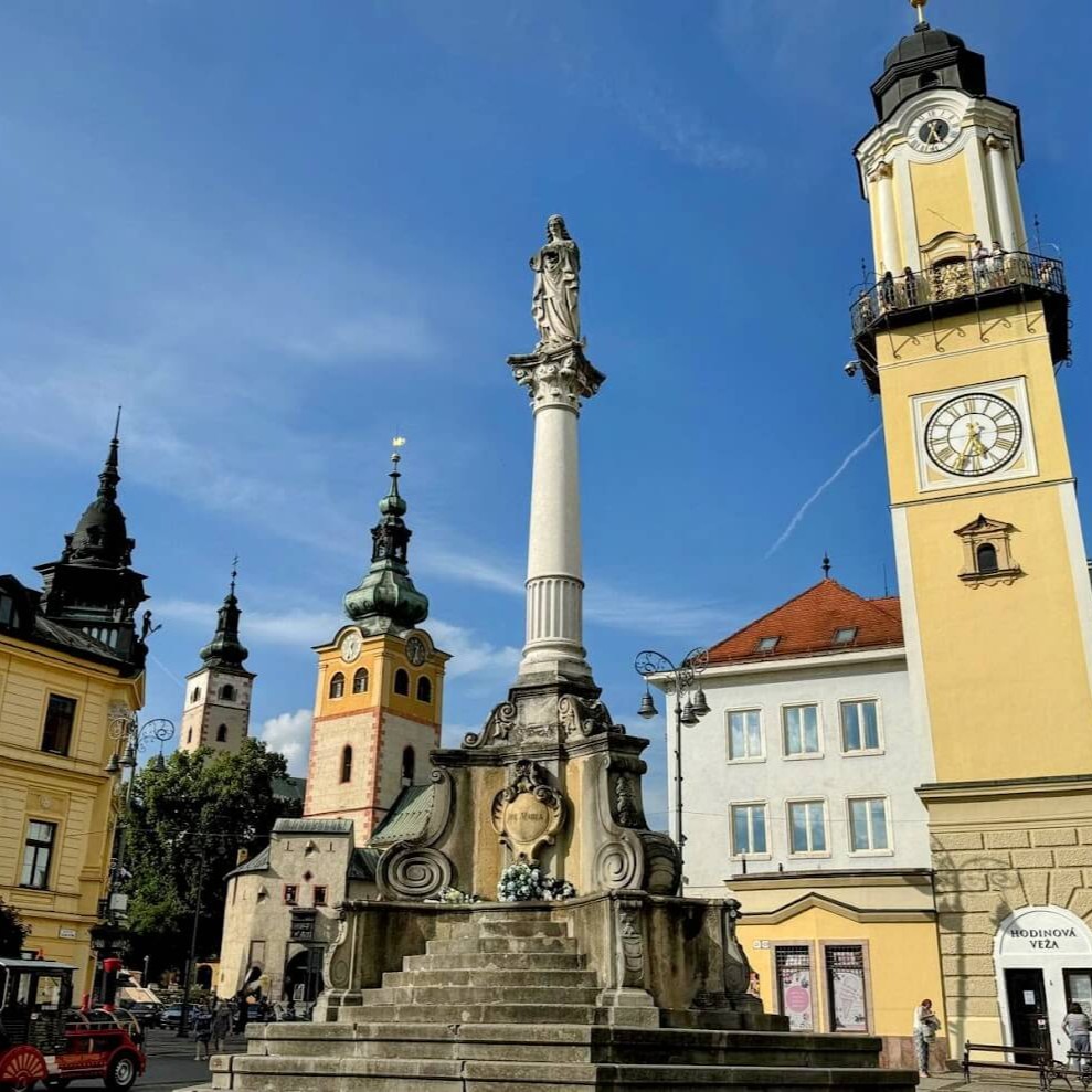 A scenic view of Banská Bystrica's historic city center featuring the Clock Tower, the Baroque Marian Column, and colorful church towers against a clear blue sky—an essential stop in any Banská Bystrica travel guide