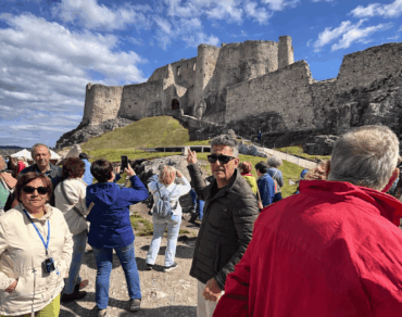 A guided tour group exploring the grounds of Spiš Castle (Spišský hrad) in Slovakia, with the massive medieval castle ruins rising dramatically on a rocky hill against a cloudy blue sky.