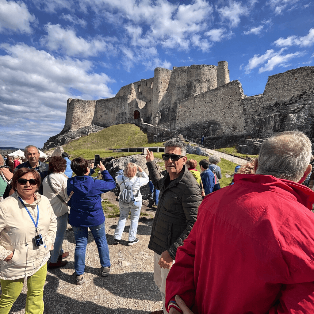 A guided tour group exploring the grounds of Spiš Castle (Spišský hrad) in Slovakia, with the massive medieval castle ruins rising dramatically on a rocky hill against a cloudy blue sky.