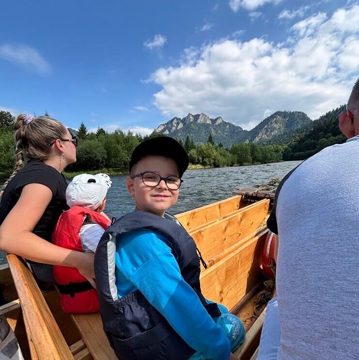 A young boy in a blue outfit and black life jacket smiles at the camera while sitting in a wooden raft on a river. Behind him are other passengers, including a woman holding a child in a red life jacket. In the background, forested hills and the distinctive Three Crowns (Tri Koruny) mountain peaks rise under a partly cloudy blue sky, indicating a scenic rafting trip in the Pieniny Mountains