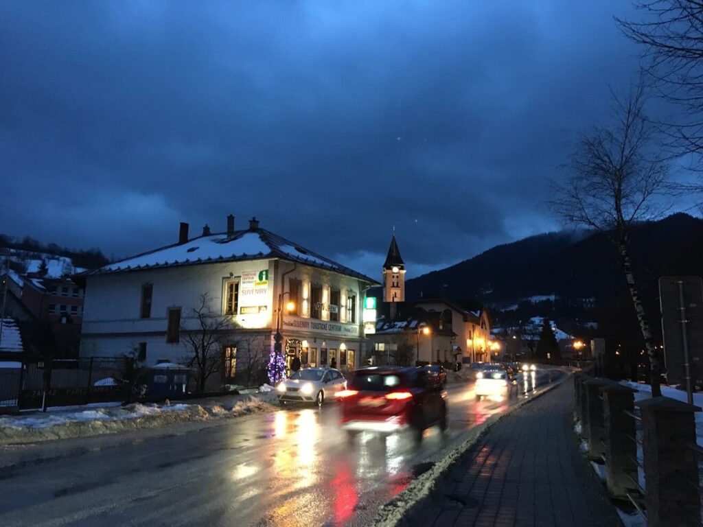 Une scène nocturne hivernale d'un village slovaque, avec des routes mouillées par la neige fondante, des lumières chaleureuses illuminant les bâtiments et une église pittoresque en arrière-plan.