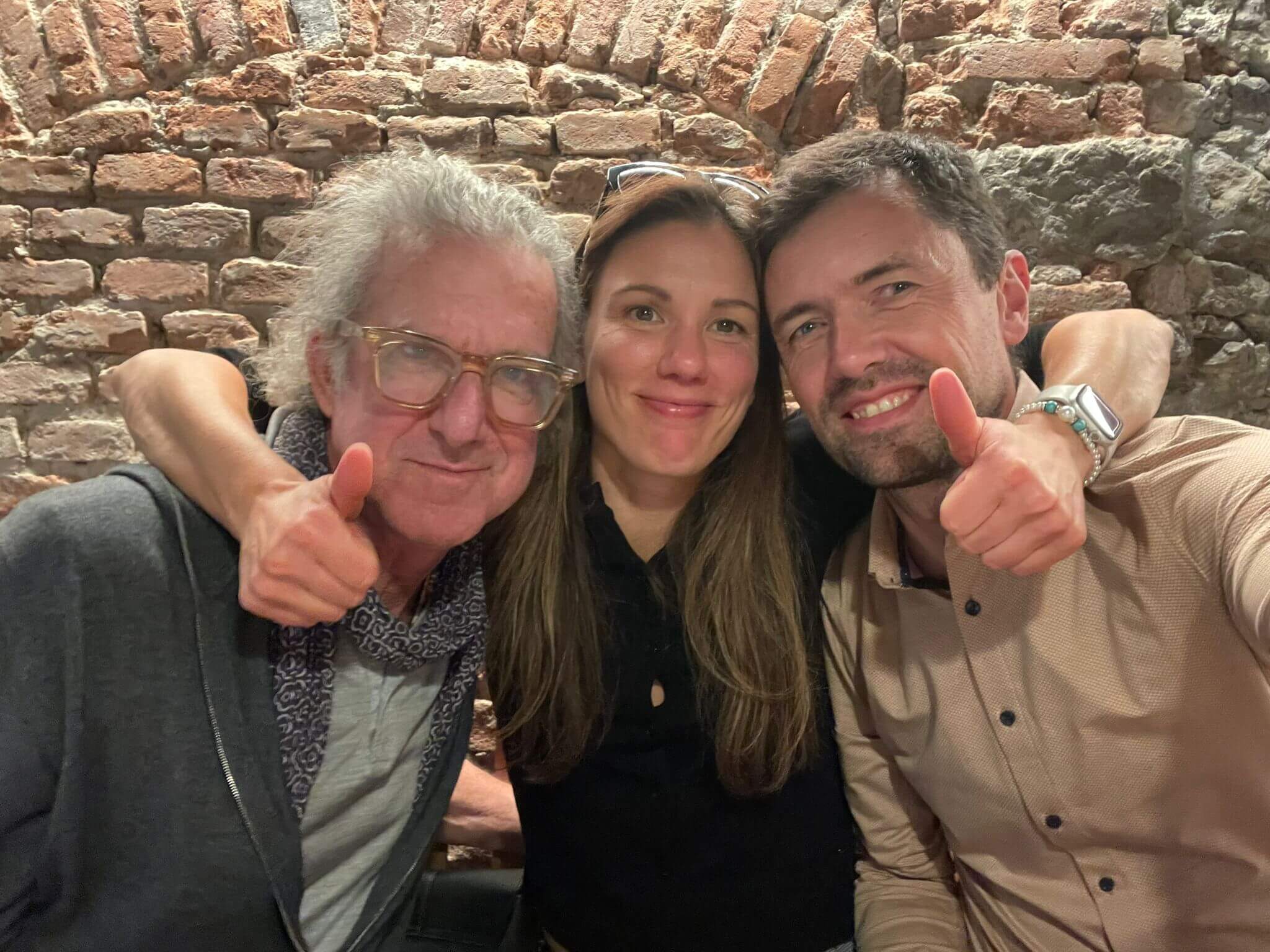 Three people smiling and giving a thumbs-up in a cozy setting with exposed brick walls, highlighting a friendly and relaxed atmosphere.