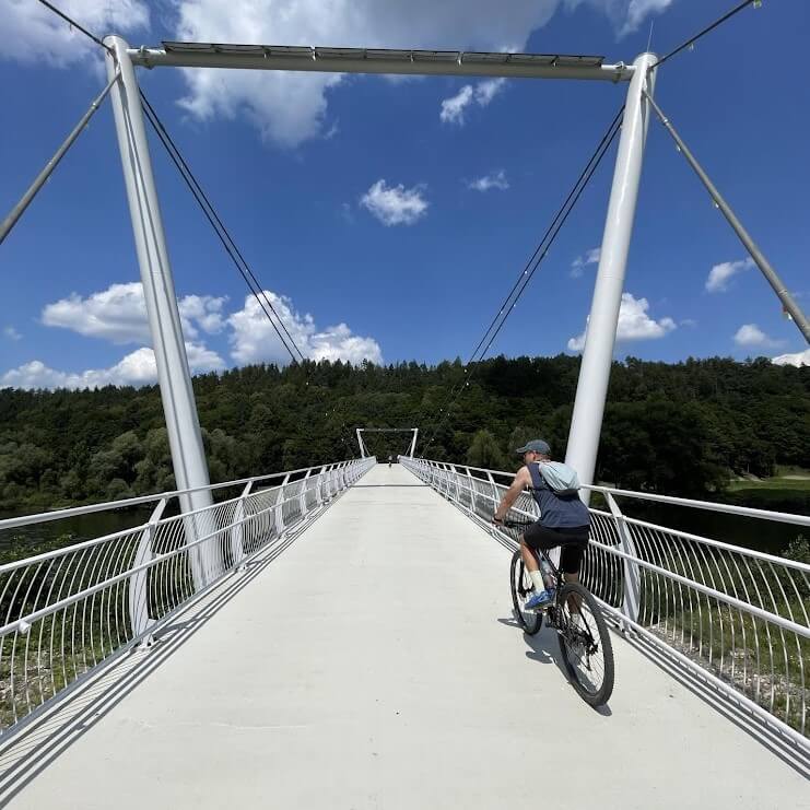 Person cycling across a modern white bridge surrounded by lush green forests under a clear blue sky, showcasing the scenic landscapes of Slovakia during a cycling program