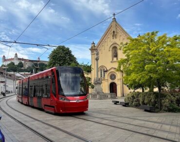 A modern red tram travels through Bratislava's historic centre, with the medieval church and Bratislava Castle visible in the background — a perfect snapshot of how the city blends old and new.