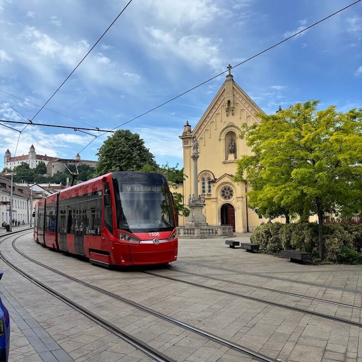A modern red tram travels through Bratislava's historic centre, with the medieval church and Bratislava Castle visible in the background — a perfect snapshot of how the city blends old and new.